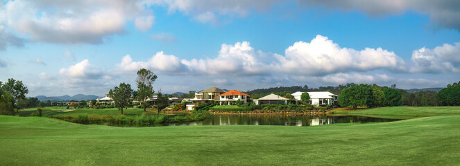 Fototapeta na wymiar Wide panoramic view of premium lush golf course with beautiful pond in the foreground, surrounded by green grass and trees. 