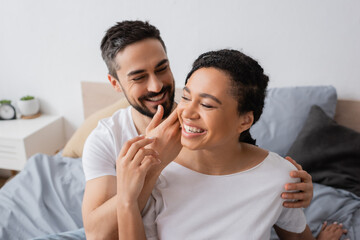 smiling bearded man touching face of overjoyed african american woman in bedroom at home.