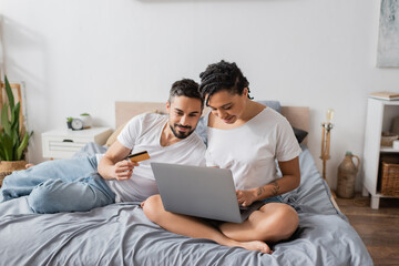 Obraz premium bearded man holding credit card near african american woman sitting with crossed legs and using laptop on bed at home.