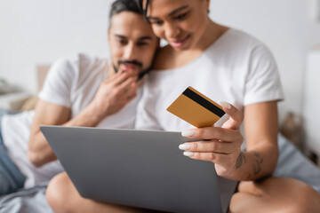 selective focus of credit card and laptop near blurred interracial couple thinking during online shopping in bedroom at home.