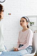 Preteen girl pouting lips during lesson with speech therapist in consulting room.