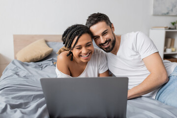 excited interracial couple laughing near laptop while watching movie on bed at home.