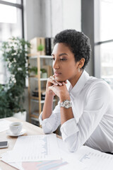 thoughtful african american businesswoman sitting near coffee cup and papers with graphs on work desk in office.