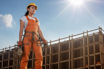 Generative AI image of young female construction worker in overall helmet gloves near site with building work and looking away in bright sun against blue sky