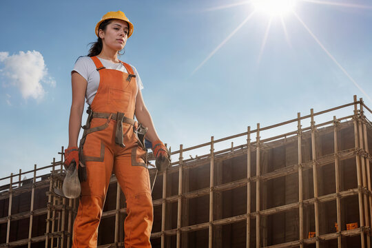 Generative AI image of young female construction worker in overall helmet gloves near site with building work and looking away in bright sun against blue sky