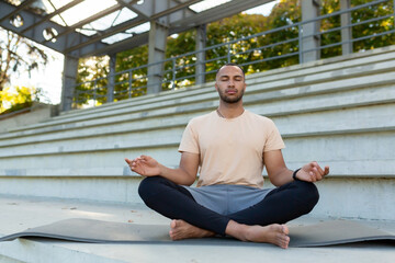 Young african american man doing yoga in stadium, urban. Sitting on a mat in the lotus position...