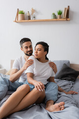 smiling man hugging african american girlfriend in white t-shirt looking away on bed at home.