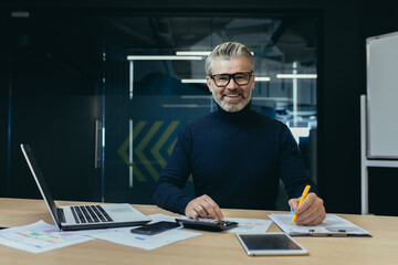 Portrait of an older gray-haired man, an accountant, an auditor sitting in the office at a laptop and a calculator. He works with documents, checks accounts. Smiling at the camera.