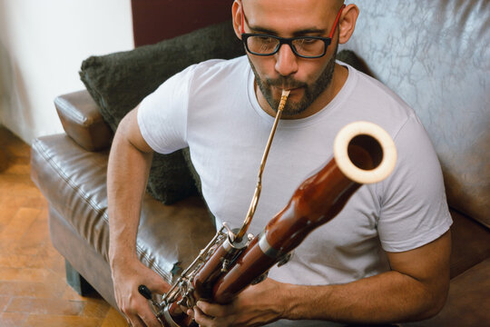 front view of latino man with beard, bald head and glasses sit at home playing bassoon.