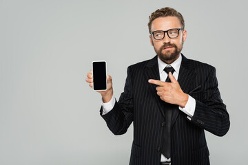 bearded businessman in suit and glasses pointing at smartphone with blank screen isolated on grey.