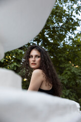 A beautiful profile portrait of a curly brunette girl in a black dress and makeup, poses gorgeous on balcony, looking at camera. 