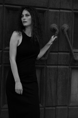 A beautiful curly brunette girl in a black dress and makeup, poses by the door. Beauty portrait. Black and white photo.