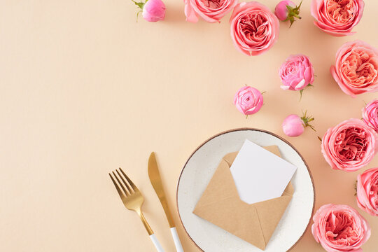 Top view photo of plate cutlery napkin with ring natural flowers pink rose buds and envelope card on light beige background with empty space. Mother's Day concept