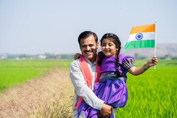Happy Indian village father carrying daughter with indian flag by walking near green farmland during independence day - concept of democracy, patriotism and relationship bonding.