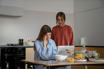 Upset depressed young woman single mother living with teen daughter sitting at kitchen table looking at laptop cant find job, mom parent struggling to pay bills, family having financial problems - obrazy, fototapety, plakaty