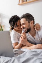 excited interracial couple laughing with closed eyes while watching comedy movie on laptop in bedroom.