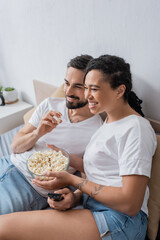 Obraz na płótnie Canvas carefree multiethnic couple in white t-shirts eating popcorn and watching tv on bed at home.