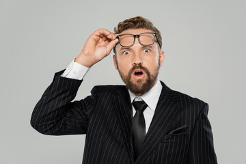 shocked businessman in formal wear adjusting glasses and looking at camera isolated on grey.