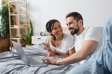 Obraz premium cheerful african american woman pointing at laptop and looking at smiling bearded man on bed at home.