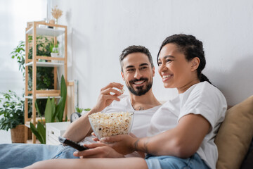 pleased bearded man eating popcorn and looking at smiling african american woman with tv remote controller on bed at home.