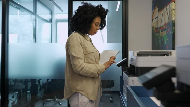 Concentrated african curly woman printing documents on printer in office
