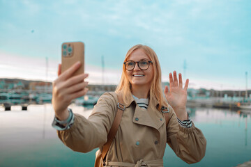 Happy 30s hipster girl blogger influencer recording vlog holding looking at camera, shooting video, streaming on pier of Sopot, Gdansk. Traveling Europe in autumn. High quality photo