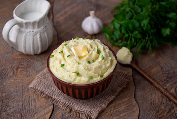 Fototapeta na wymiar Garlic Mashed Potatoes. Creamy Buttery Mashed potato topped with a knob of butter and chopped green scallion. Rustic brown bowl on wooden table with bunch of parsley and jug of milk on background.