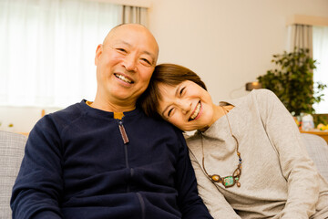 自宅のリビングで肩を預けて甘えるとても仲良しのカメラ目線の50代から60代のシニアの夫婦の笑顔