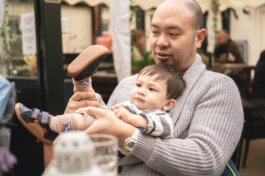 Flexible male infant sitting on his dad's legs, holding his own legs, playing with father, serious expression, in beer garden at pub in Scotland. Happy moment. Multicultural family, mixed race.
