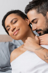 charming african american woman smiling with closed eyes while bearded man hugging her in bedroom at home.