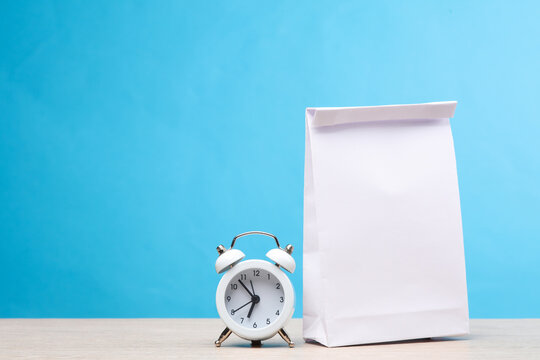 White lunch bag with alarm clock on the table, blue background. School time