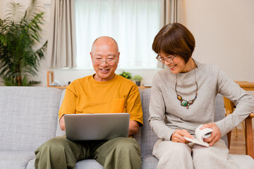 自宅のリビングでソファに座って膝の上のノートパソコンを操作する男性と読書をする女性の老夫婦