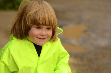 A small cute boy with long hair in a light green raincoat with a hood and rubber boots. The child jumps through the puddles, the boy rejoices in the rain, spring, autumn.