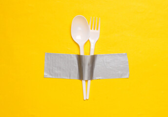 Plastic fork and spoon fixed with adhesive tape on yellow background. Conceptual pop, contemporary art, minimal food concept