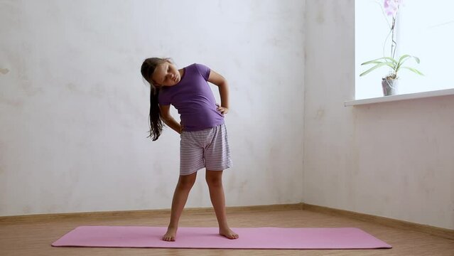 School age girl in sportswear standing and sluggishly and too slow bending body sideways on gymnastic mat in empty room, free copy space. Home workout, gymnastics, kids sport, exercise, activity.