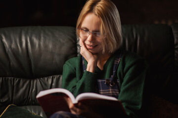 A woman with glasses reads a book on the sofa at home. Forty years old woman is reading a fascinating book.