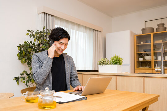 自宅でノートパソコンを使ってテレワーク・在宅勤務中にスマホで電話をする30代の男性