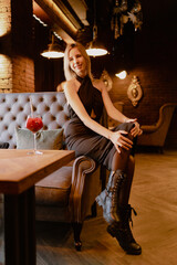 Portrait of young joyful perfect woman wearing long black dress, boots with fair hair sitting with crossed legs on arm of brown sofa in restaurant near glass with red cocktail with straw. Vertical.