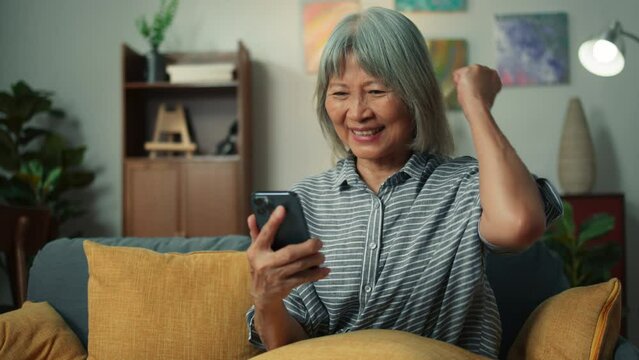 Happy Asian elderly woman smile holding mobile phone checking email received good news, feeling surprised excited. Cheerful aged female looking at smartphone screen sit on sofa in home living room.