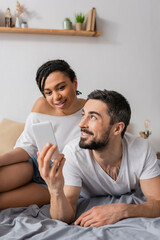 cheerful bearded man showing smartphone to smiling african american woman while resting on bed at home.