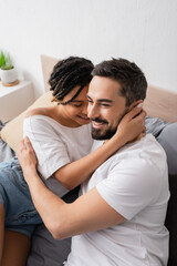 young african american woman and happy bearded man in white t-shirts embracing in bedroom at home.