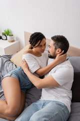 high angle view of multiracial couple in white t-shirts embracing and looking at each other on bed at home.