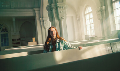 A beautiful young red-haired woman is sitting on old pews inside a Protestant church, illuminated...