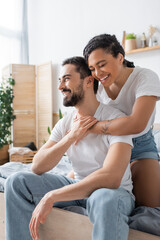 joyful african american woman with closed eyes hugging happy boyfriend in white t-shirt and jeans sitting on bed at home.