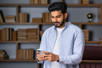 Indian man holding smartphone using mobile phone at home looking at cell reading mobile new...
