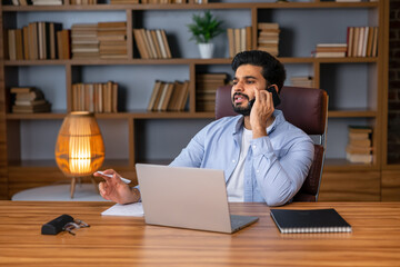 Fototapeta na wymiar Handsome arab man with beard sits at work desk with laptop, talk by phone, looks at empty space in living room interior. Work at home, social networks, call to client remote, great offer and ad