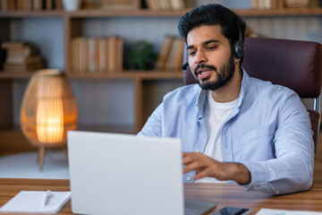 Fototapeta na wymiar Young bearded man using laptop computer during video call working in office. Concentrated adult successful man wearing casual clothes sitting at wooden desk indoor.