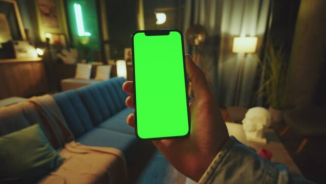 Hand man holds a scrolling phone with a vertical green screen in a dark room. Device. Smartphone. Social media
