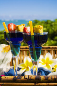 Two Tropical Drinks Garnished With Flowers And Fruit In An Outdoor Setting.