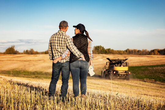A view taken from behind of a husband and wife standing in a field spending some quality time together while watching a combine finish their fall canola harvest; Alcomdale, Alberta, Canada
